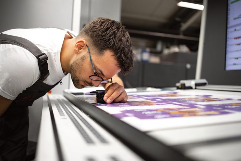 A print technician at Master Printing + Mailing using a magnifying loupe to inspect color consistency on a press sheet, illustrating G7 Master Targeted quality standards.